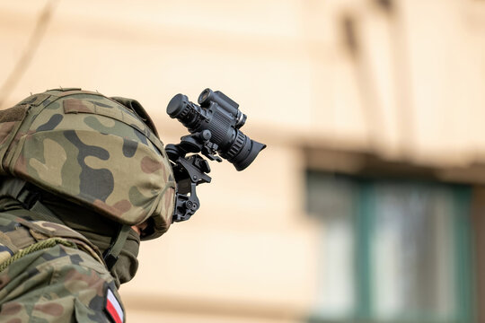 Polish infantry soldier with helmet-mounted night vision goggles, camouflaged uniform and face mask, side view closeup against blurred urban background