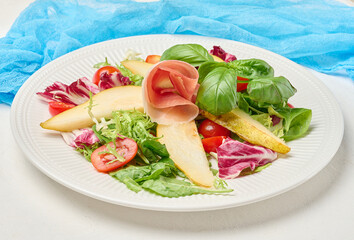 Close up of a gourmet salad with prosciutto, pear slices, cherry tomatoes and fresh mixed greens served on a white ribbed plate.