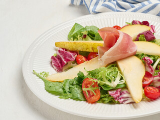 Close up of a gourmet salad with prosciutto, pear slices, cherry tomatoes and fresh mixed greens served on a white ribbed plate.