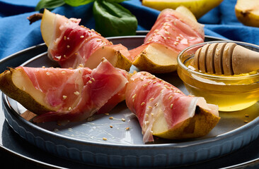 Gourmet appetizer of pear with prosciutto and sesame seeds, served with honey on a blue ceramic plate.