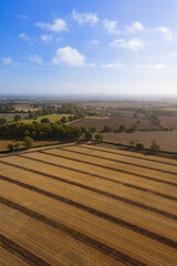 Aerial view of the british countryside during sunrise, with freshly harvested, tree lined, agricultural farm fields. Bright blue sky with sunburst and white fluffy clouds. Vertical panorama