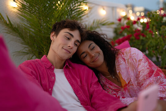 Young couple sit together on couch, smiling and looking at a smartphone in a cozy outdoor setting.