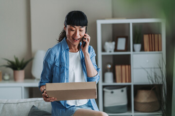 Happy asian woman opening parcel and talking on mobile phone at home