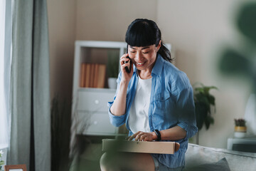 Happy asian woman opening parcel and talking on mobile phone at home