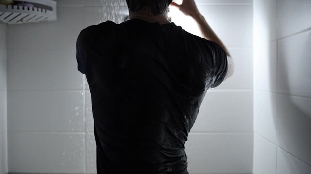 Young man in wet black shirt stands under running shower reaching up with both hands as if fixing the shower head while water pours down around him