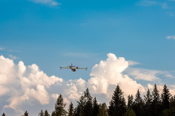 Yellow firefighting plane approaching forest under bright sky.