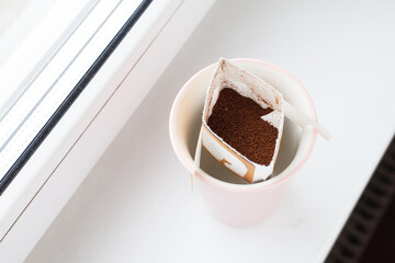 Drip bag in a ceramic cup on windowsill. Instant freshly brewed alternative filter drink in mug. Easy brew black coffee method for home, travel or office. cozy trendy lifestyle