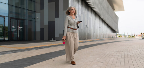 Mature woman walking confidently on modern plaza in urban setting during daytime while enjoying coffee