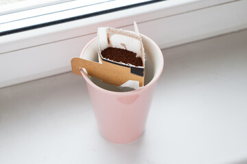 Drip bag in a ceramic cup on windowsill. Instant freshly brewed alternative filter drink in mug. Easy brew black coffee method for home, travel or office. cozy trendy lifestyle