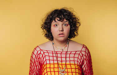 A person with curly hair wearing a red net top over a yellow shirt, standing against a yellow background.