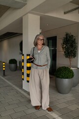 Senior woman in stylish outfit stands near potted plants in urban setting during daytime