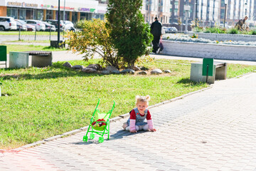 A little caucasian girl on the sidewalk, with a green toy baby stroller nearby on a sunny day in...