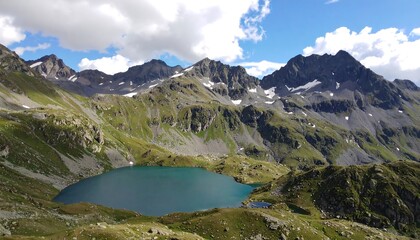A high-elevation alpine lake nestled amongst rugged, snow-capped mountains under a partly cloudy sky. Lush green meadows and gray rock formations frame the serene body of water.