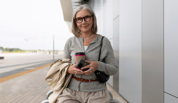 Senior woman holding a coffee cup and smartphone while waiting at an airport terminal on a cloudy day