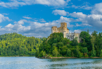 Medieval Niedzica Castle stands on a forested hill above Czorsztyn Lake, reflected in blue water....