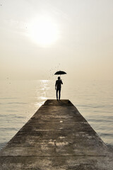 Fototapeta premium A man in silhouette, holding a black umbrella, walks on a concrete pier towards the sea. The landscape is dominated by a golden, foggy light. The image is suitable for concepts of solitude