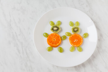 Creative healthy snack of breakfast for kids fruit bunny shaped on white plate, easter eggs around. Rabbit, basket makes from grape, orange, banana, apple. Top view