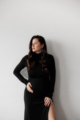 Naklejka premium Pregnant woman in a black dress posing against a white wall, smiling and holding her belly.