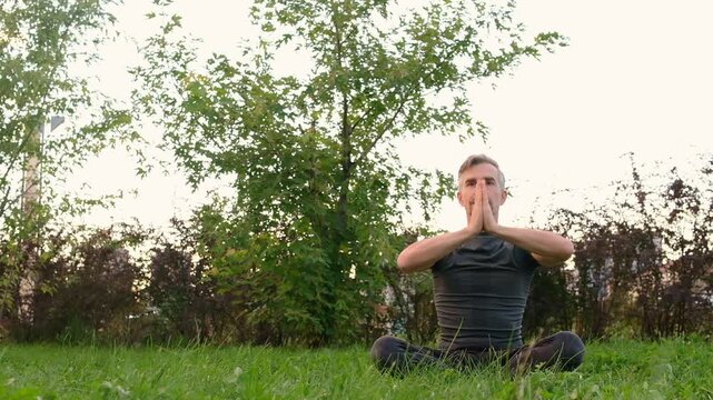 Mature man practicing yoga sitting in lotus pose on green grass in park outdoors