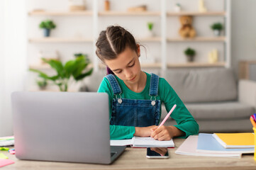 Front portrait of focused girl sitting at table with laptop computer in living room at home, writing essay or making test exam, taking notes in textbook, doing homework, free copy space