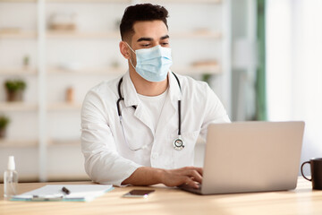 Arab doctor in protective face mask using laptop at clinic, typing on notebook keyboard. Middle-eastern physician giving online consultation during COVID-19 pandemic, remote medicine concept