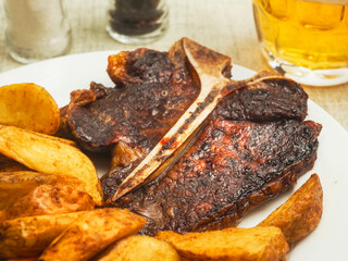 Meal with high quality T bone steak, wedges, fresh tomatoes and pickles, glass of lager beer and pepper and salt shakers. Top version of beef strip loin with bone in for a special great flavor.