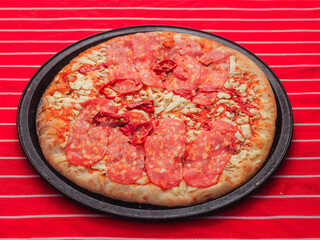 A meat feast pizza with pepperoni, ham and cheese on it. The pizza is on a black plate and is sitting on a red tablecloth. Premium high quality ingredients. Italian style food.