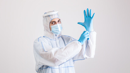 Young male virologist in hazmat suit putting on rubber gloves on light studio background. Doctor wearing protective biohazard costume while fighting coronavirus. Viral diseases control concept