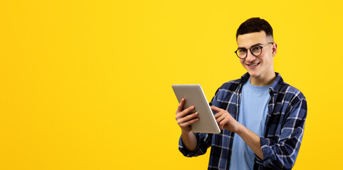 Online life concept. Funky young man using tablet pc on orange studio background. Joyful millennial guy with touch pad surfing web, watching videos, checking email, posting in social media