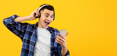 Excited Caucasian guy in wireless headphones using smartphone on orange studio background. Funky...