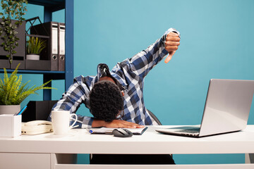 Black businessman showing dislike gesture and resting his head on office desk, struggling to stay...