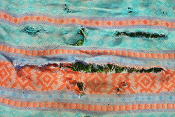 texture of old torn green with orange patterns fabric with horizontal long holes and uneven edges lies on the grass