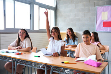 Group of teenage students sitting at desks in a classroom during a lesson, with one girl raising her hand eagerly to answer a question posed by the teacher