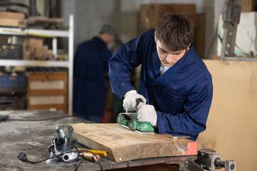 Young guy worker in carpentry workshop polishes wooden board with machine