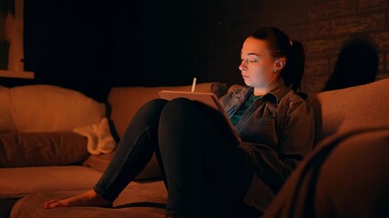 Focused Caucasian woman working from home sitting on a sofa while using a digital tablet and stylus to create detailed drawings. Freelance designer is deeply concentrated on sketching and illustrating - Powered by Adobe