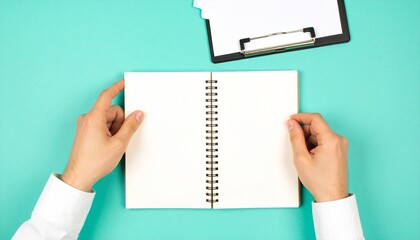 Obraz premium Hands holding open blank spiral notebook on a vibrant teal background with a clipboard, offering copy space.