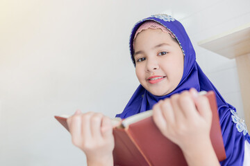 Young Muslim girl in blue hijab smiling while reading a book, studying at home with white background. Education and learning concept for Islamic students.