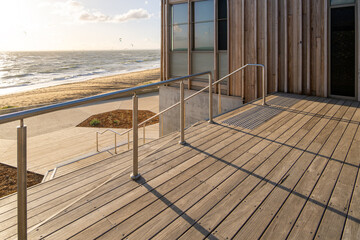 Modern outdoor plaza with wooden decking and stainless steel railing at beach, featuring community space and stairs providing direct access to the sandy beach. Coastal architecture, leisure amenities