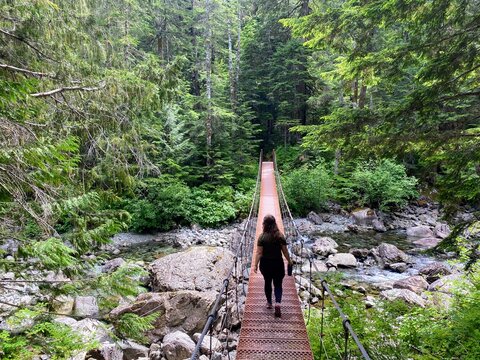 Woman hiking across a narrow suspension bridge over a rocky creek on Bedwell Lake Trail in Strathcona Provincial Park, Vancouver Island, BC forest.