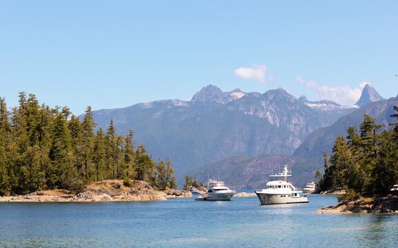 Luxury yachts anchored in a calm blue inlet surrounded by evergreen trees and rugged mountains under a clear summer sky at Prideaux Haven, Desolation Sound, British Columbia.