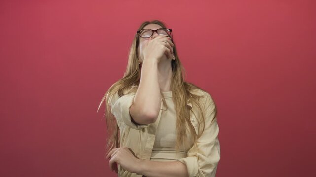 Young blonde woman with long hair and glasses covers mouth with hand while tilting head in studio; amusement.