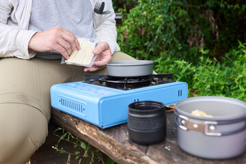 Camping meal: an unrecognizable person unwraps a couple of sandwiches to toast them in a pan using a portable gas stove.