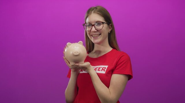 Young blonde woman wearing volunteer shirt smiling holds piggy bank in studio; generosity charity donation.