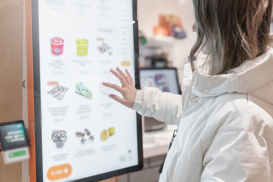 A woman is interacting with a digital menu displayed at a modern retail store today. A 30 -35-year-old woman in a white winter jacket orders food through the screen menu at a fast food restaurant