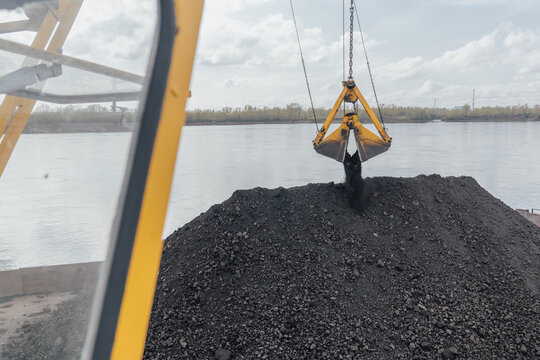 Coal loading by crane onto a barge on the river, northern delivery along the Yenisei in Russia. View from the crane cabin. Coal bucket loading and unloading process at a loading dock by an excavator