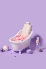 Composition with mini bathtub, bottles of cosmetic products and bomb on lilac background
