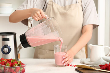 Woman pouring tasty strawberry smoothie from blender into glass at table in kitchen