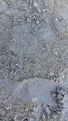 Coarse, grey sand covers a surface with a blend of dark and light particles, indicative of volcanic origin, typical of certain Balinese beaches. The texture appears gritty, with scattered small stones