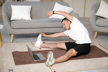 Naklejka premium Sporty young man with laptop stretching on mat at home
