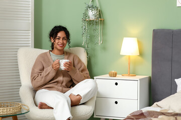 Beautiful young happy African-American woman in stylish outfit with cup of tea sitting on armchair...